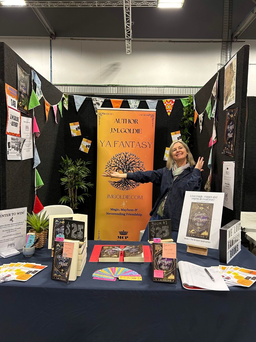 J.M. Goldie at her book stall at Armageddon Expo, books laid out on the table in front of her YA fantasy banner